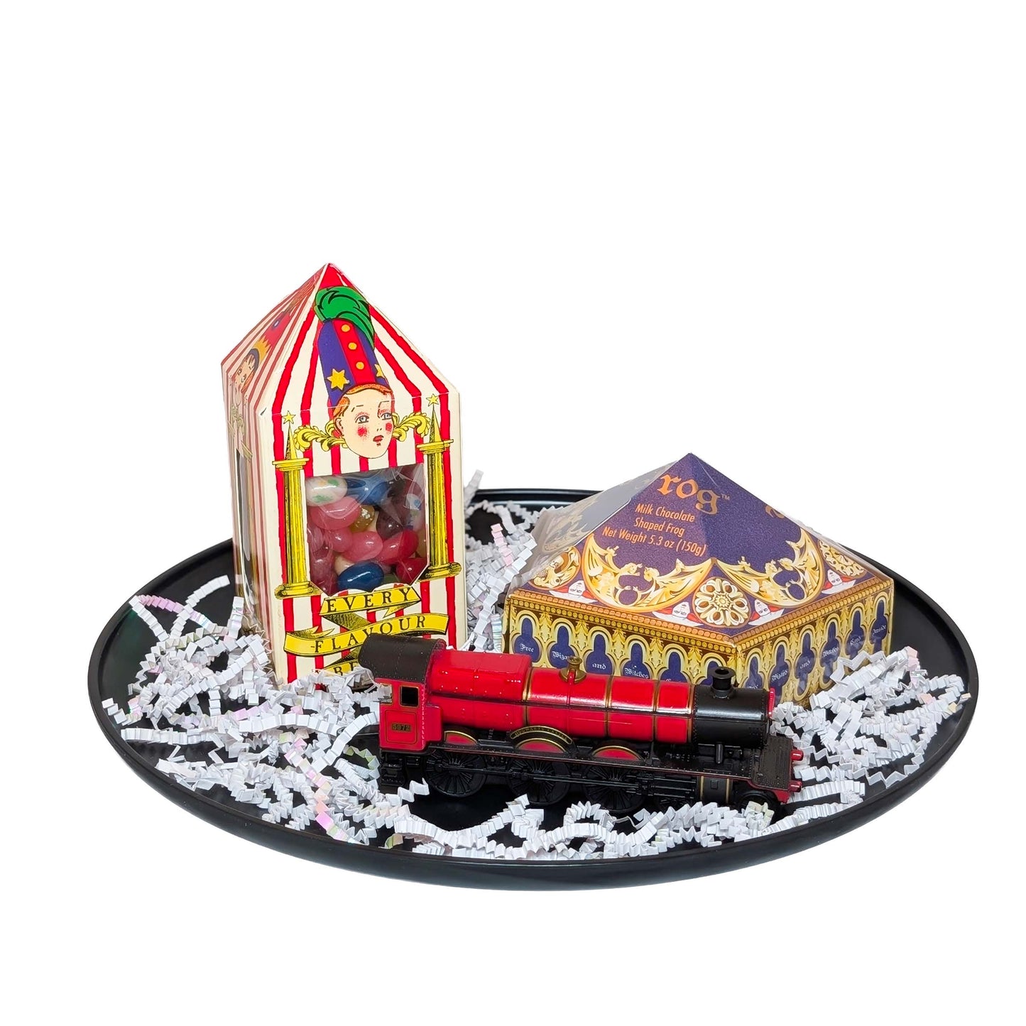 Hogwarts Express toy train, a Wizarding World Chocolate Frog box, and Bertie Bott's Every Flavor Beans box arranged on a matte black tray with white shredded paper fill for an Orlando resort gift basket