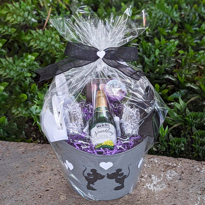 A gift basket containing a bottle of sparkling grape juice, two stemless wine glasses, a box of Disney chocolates, and a selection of Disney park snacks, elegantly wrapped with a bow and transparent film, perfect for deliveries at Walt Disney World