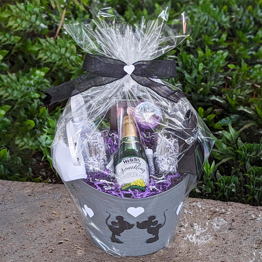 A gift basket containing a bottle of sparkling grape juice, two stemless wine glasses, a box of Disney chocolates, and a selection of Disney park snacks, elegantly wrapped with a bow and transparent film, perfect for deliveries at Walt Disney World