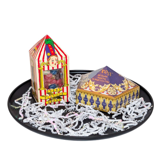 A Wizarding World Chocolate Frog box and Bertie Bott's Every Flavor Beans box arranged on a matte black tray with white shredded paper fill for an Orlando resort gift basket