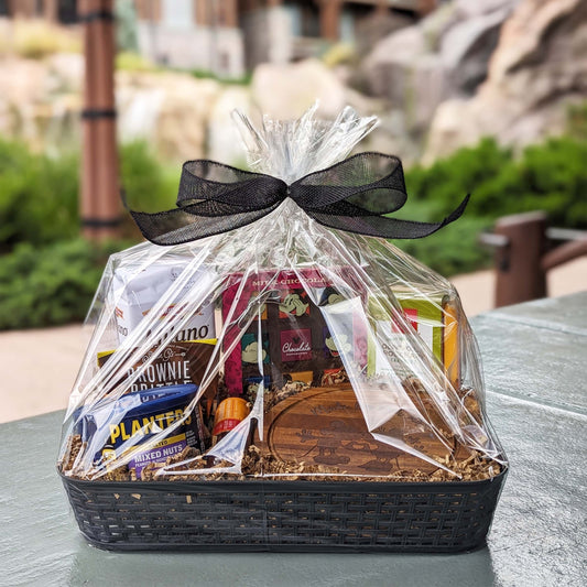 Charcuterie board gift basket wrapped in clear plastic with a black ribbon, featuring an assortment of meats, cheeses, cookies, brownie brittle, chocolate candy, and nuts